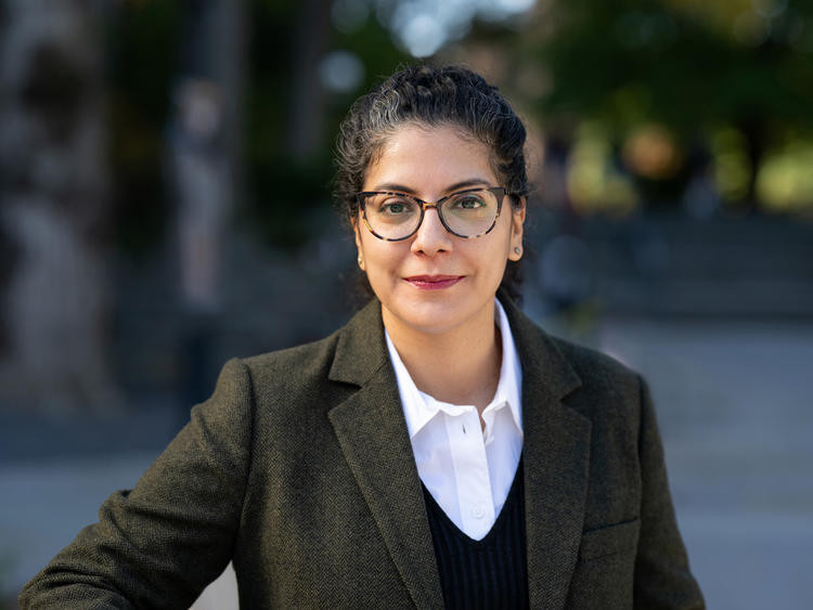 A confident woman with glasses, in a dark blazer and white shirt, stands outdoors. The blurred background features greenery and soft lighting.