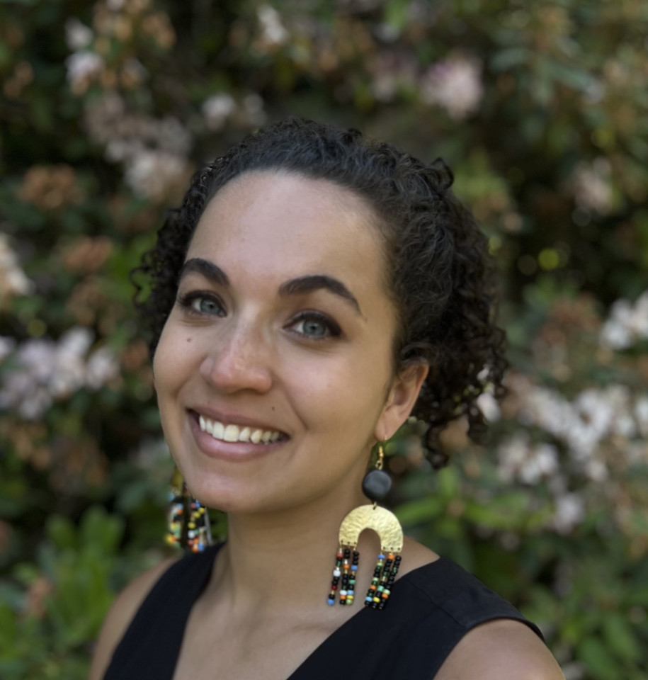 Smiling person with curly hair, wearing colorful beaded earrings, stands in front of blurred greenery and pink flowers, exuding a warm, joyful vibe.