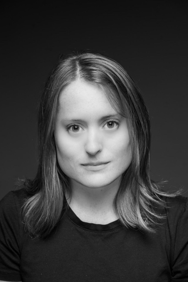 A black-and-white portrait of a person with shoulder-length hair, wearing a black shirt, looking directly at the camera with a calm expression.