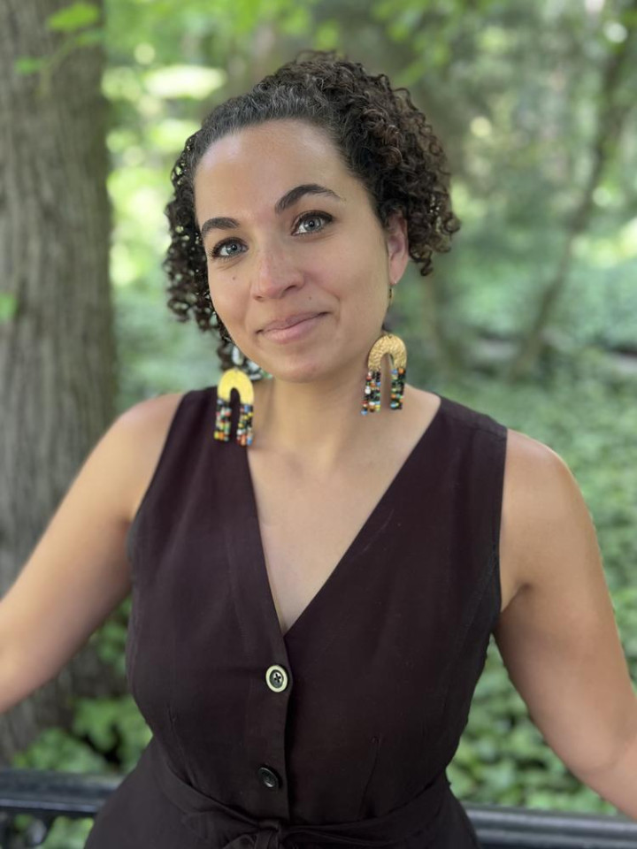 A woman with curly hair stands outdoors near trees, wearing a sleeveless dark dress and colorful, dangling earrings. She smiles softly in the serene setting.