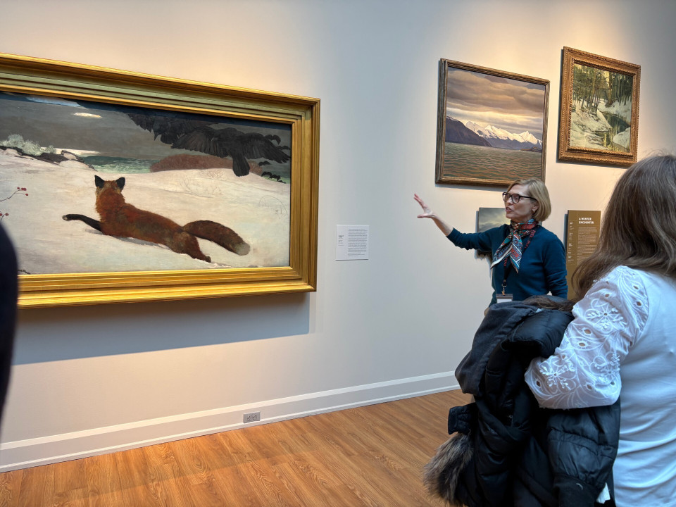A woman gestures toward a large painting of a fox in snow in a museum gallery. Several other landscape paintings hang on the wall. Visitors listen attentively.
