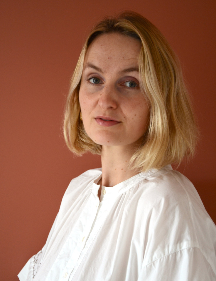 A person with short blonde hair and a white shirt stands in front of a plain, muted red background, looking slightly to the side of the camera.