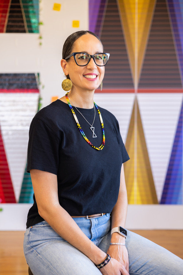 A smiling woman with dark hair tied back sits against a colorful geometric background, wearing a black t-shirt, gold earrings, and a colorful beaded necklace.