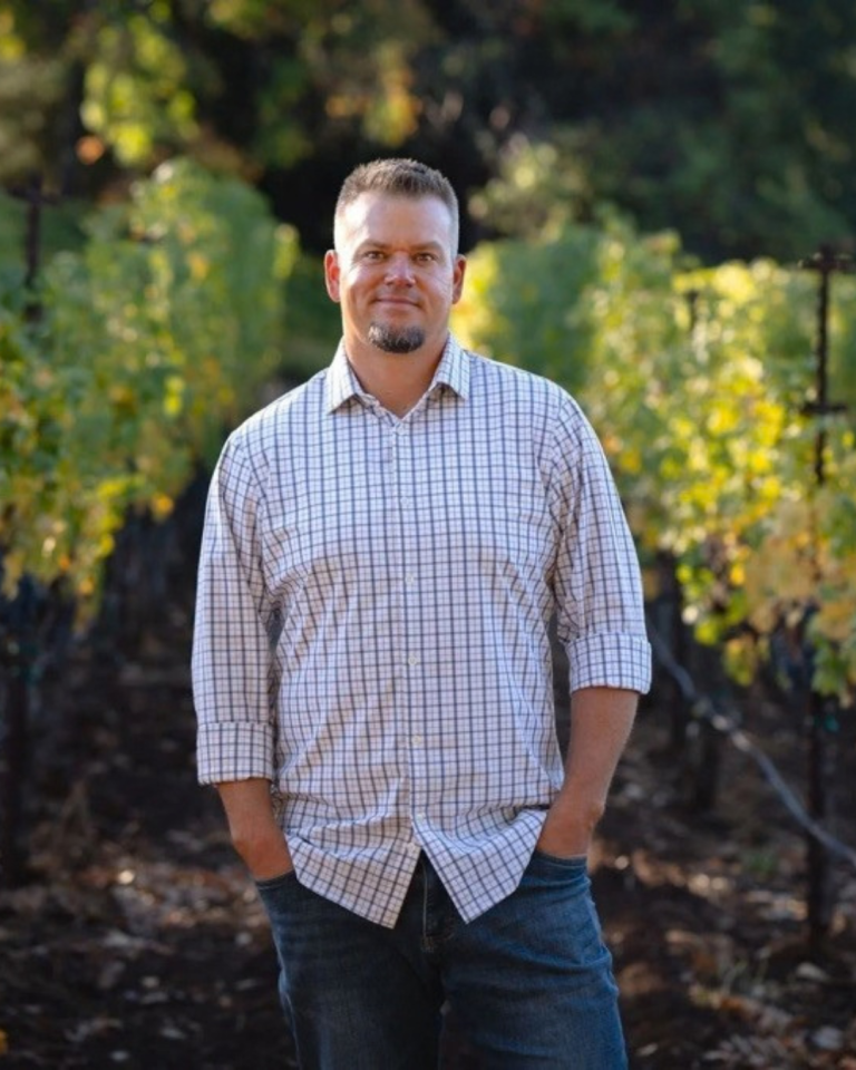 A medium shot of a man with a short beard and haircut, wearing a plaid button-down shirt and blue jeans, standing with his hands in his pockets in a sunlit vineyard.