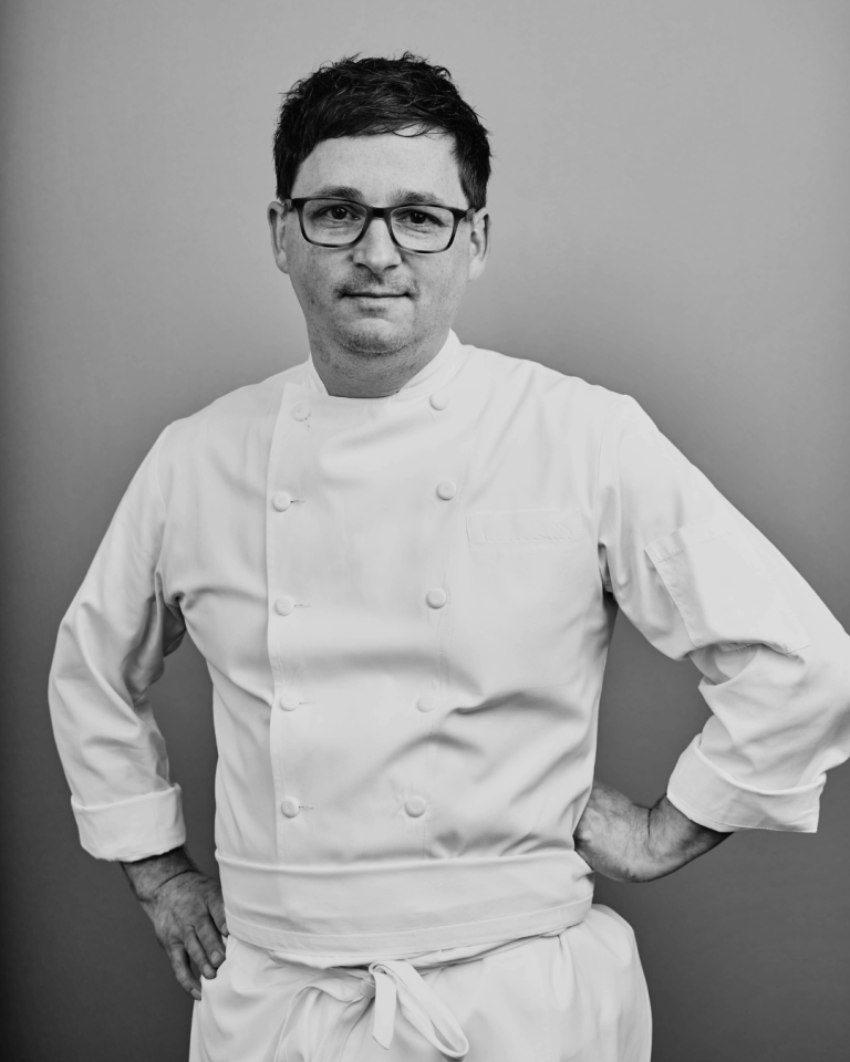 A black and white portrait of chef Nicholas Bazik wearing a double-breasted white chef's jacket and glasses, standing with his hands on his hips against a plain background.