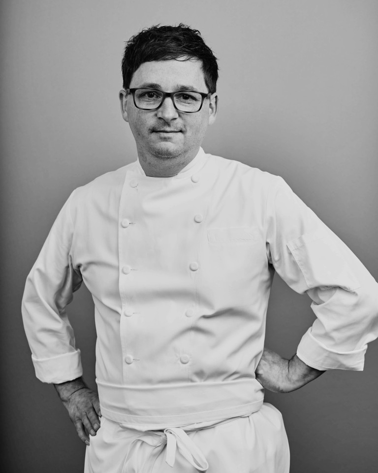A black and white portrait of a man with glasses wearing a white double-breasted chef's uniform standing with his hands on his hips against a plain background.