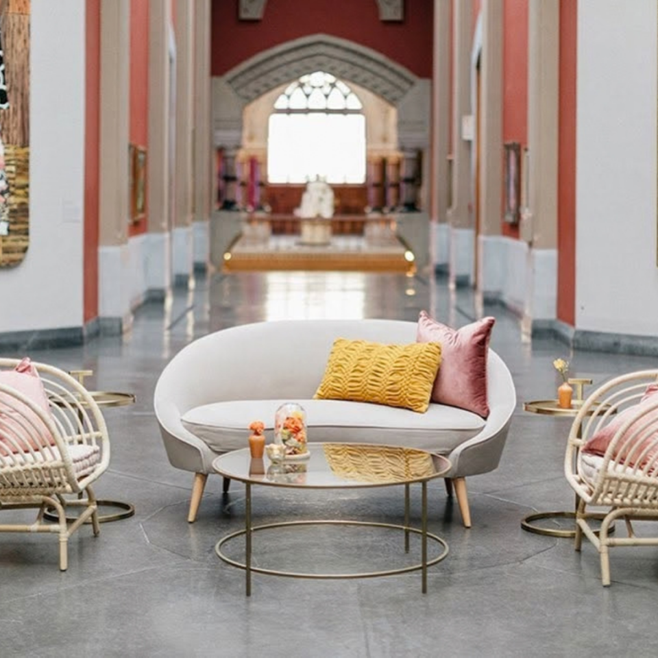 A stylish indoor seating arrangement features a curved cream loveseat paired with a round glass coffee table, flanked by two matching rattan accent chairs on a sleek grey floor within a grand, high-ceilinged hallway.