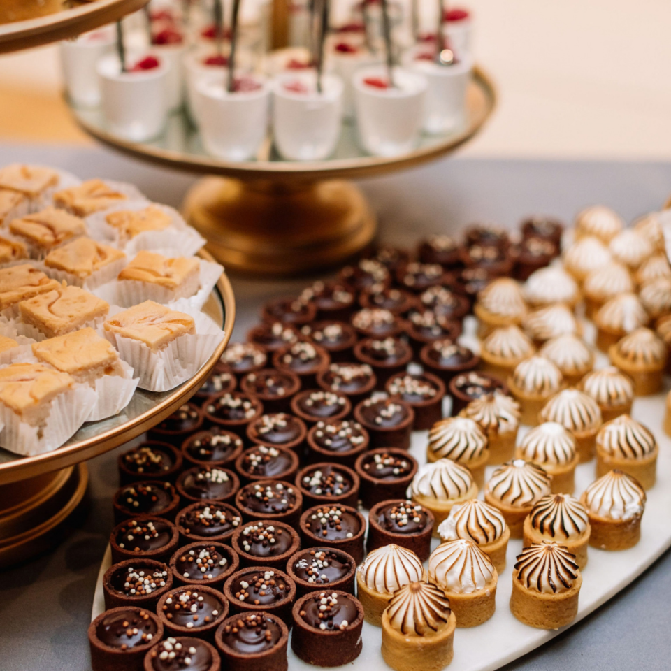 A neatly arranged dessert spread features rows of miniature dark chocolate tarts, small meringue-topped pastries, and square-cut blonde bar desserts on tiered displays.
