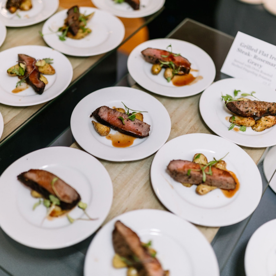 Multiple rows of individually plated steak dinners are arranged neatly across a catering station at a catered event.