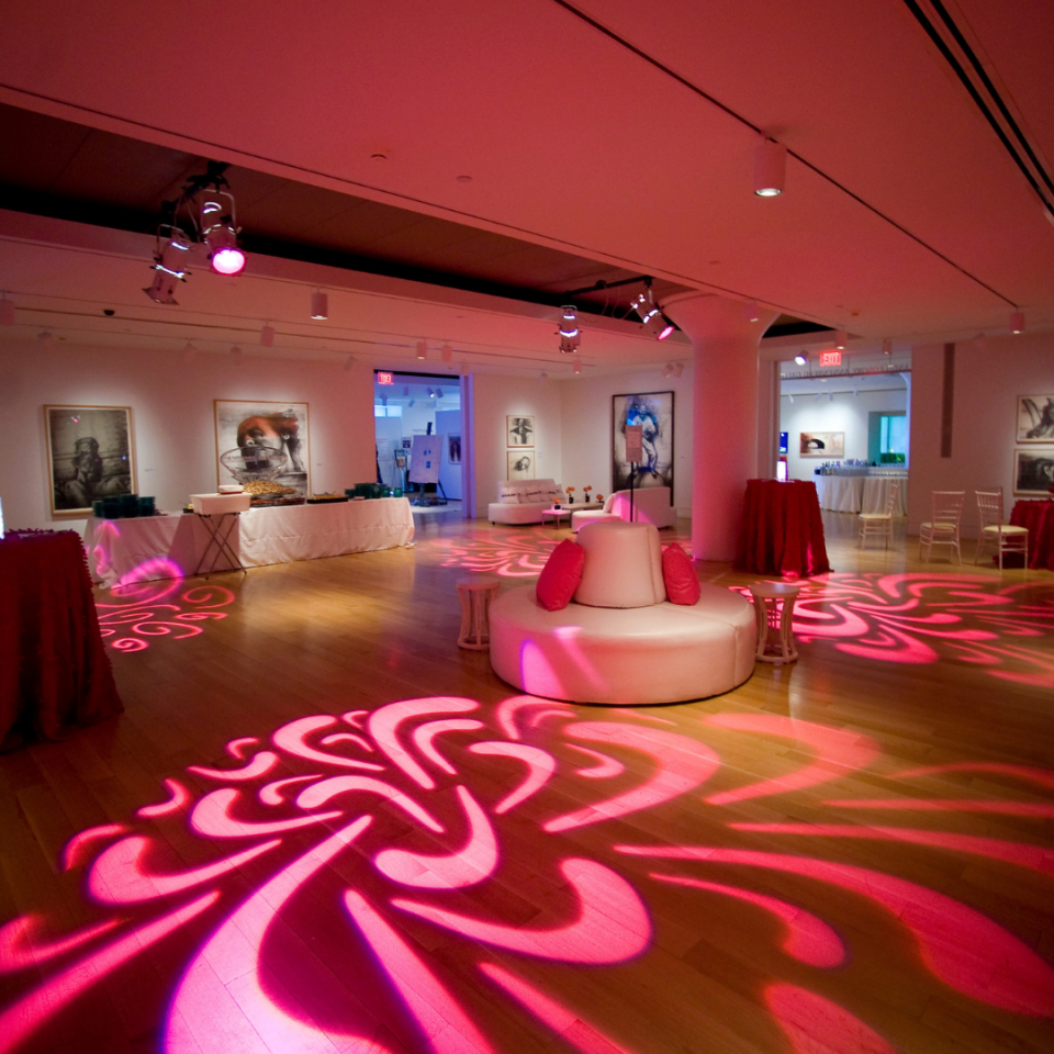 An event venue with light wood floors features a large circular white lounge sofa with pink pillows, surrounded by dynamic pink patterned lighting projected onto the ground.