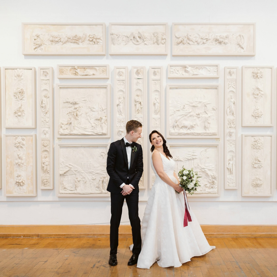 A newlywed couple smiles at each other while posing in front of a gallery wall covered in white, framed plaster relief sculptures.