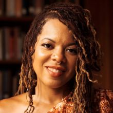 Bio photo of Airea D. Matthews a brown-skinned person, with long wavy brown hair, smiling and tilting their head to the right (a bit) 