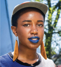 Head and shoulders bio photo of Petra Floyd, a brown-skinned person with short-cropped hair and wearing blue lipstick, long wooden earrings, and a white baseball cap. 