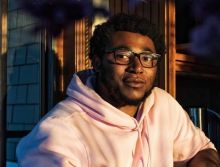 Bio photo of TK Smith, a dark-skinned man with a chin beard, wearing black-framed glasses and a white hoodie. He looks like he is sitting in afternoon sunlight. 