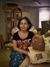Photo of Kukuli Velarde, a light brown skinned woman with dark wavy hair cut above the shoulders, sitting in a studio, left of a piece of red clay on a table, being formed into a vessel-like object. 