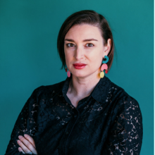 Bio photo of Leah Triplett Harrington, a light skinned woman with dark, straight hair cut below the ears, colorfull earrings, and a black blouse. 