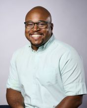 bio photo of Marvin Edward, a person with brown skin, bald head and wearing glasses. Marvin is posed seated in a traditional portrait pose wearing a mint blue button down short-sleeved shirt. 