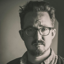 A black and white close-up portrait of the filmmaker Evan Goodchild, who is wearing dark-rimmed glasses and looking at the camera.