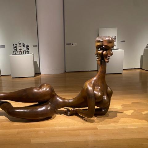 View of a wooden sculpture on display in an art gallery with a person in the background.