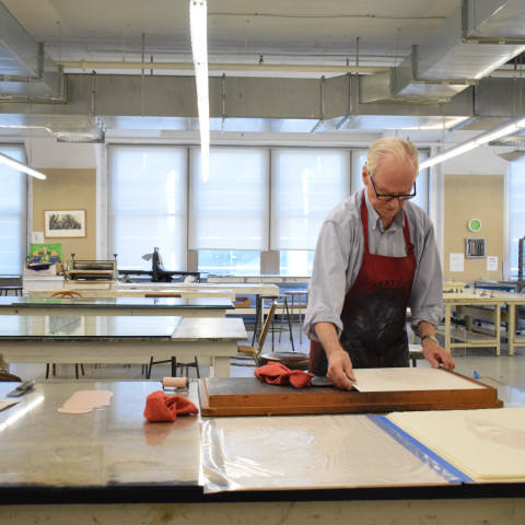 An older man wearing glasses and a red apron carefully aligns a sheet of paper over a printmaking block inside a brightly lit art studio.