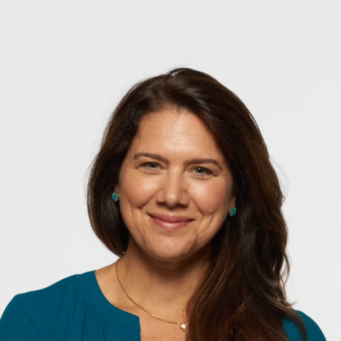 A headshot of a smiling woman with long brown hair, wearing a teal blouse, a thin gold necklace, and small teal stud earrings against a plain white background.