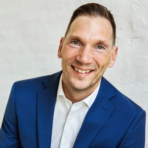A smiling man with short brown hair is wearing a bright blue suit jacket over a crisp white collared shirt against a white brick wall.