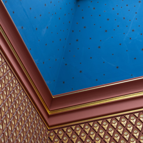 A high-angle shot captures an ornate corner ceiling featuring a bright blue panel dotted with small gold stars, framed by thick reddish-brown and gold trim above walls covered in geometric gold and brown patterns.