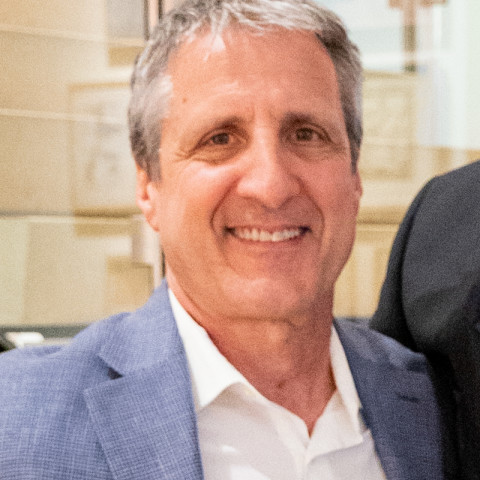 A close-up portrait of a smiling man, wearing a blue blazer and a white collared shirt, facing the camera.