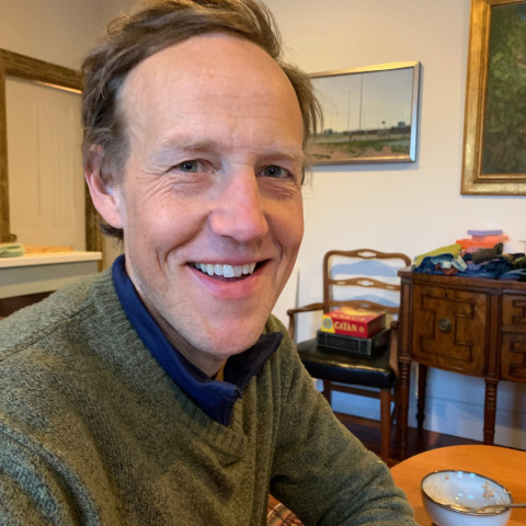 A smiling man wearing an olive green sweater sits at a dining table with a bowl, in a dining room featuring a Catan board game on a chair and framed artwork on the wall.