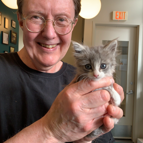 A smiling man with glasses and a black shirt gently holds a small, fluffy gray and white kitten in both hands inside a room with an exit sign.