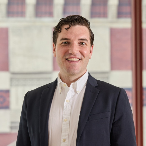A smiling man with wavy dark hair wears a navy blue blazer over an unbuttoned white shirt, posing for a professional headshot in front of a window.