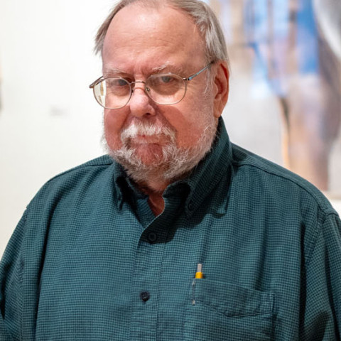 A medium close-up of a fair-skinned older man with glasses, a white mustache, and a grey beard wearing a green button-down shirt over a dark undershirt, positioned against a blurred background.
