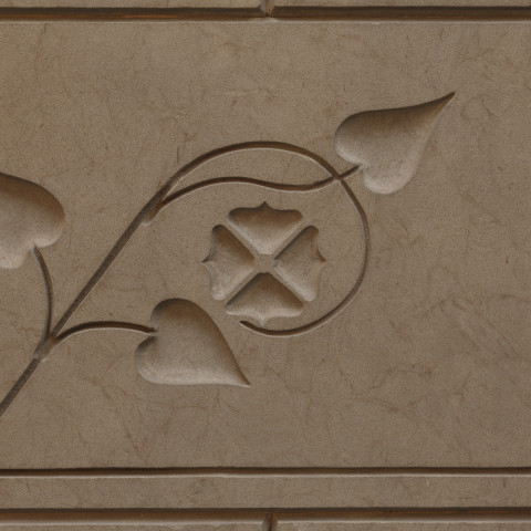 A rectangular stone panel features a low-relief architectural carving of a stylized vine with three heart-shaped leaves curving gracefully around a central quatrefoil flower emblem.