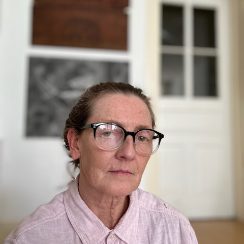 A close-up portrait shows a woman with glasses and her hair tied back, wearing a light pink button-down shirt against a blurred interior background featuring framed artwork.
