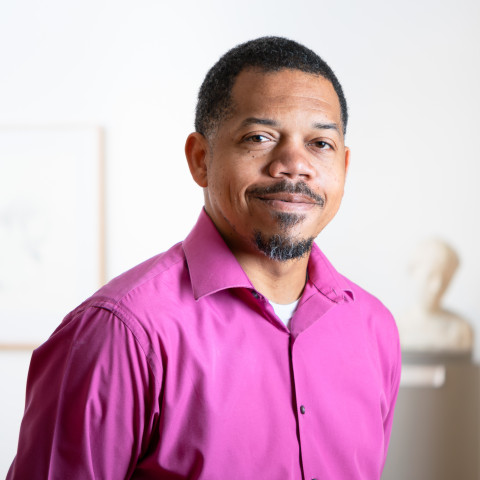 A medium shot of a Black man with a goatee and short dark hair smiling, wearing a vibrant magenta button-down shirt.