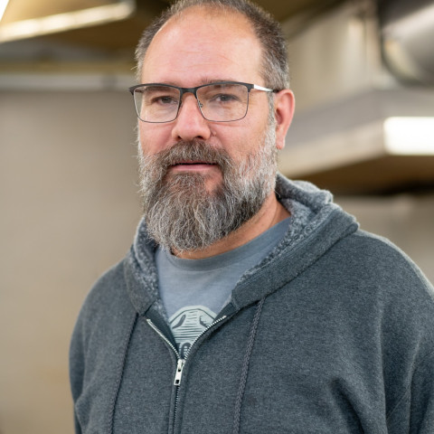 A medium close-up shot shows a middle-aged man with glasses and a grey beard wearing a charcoal grey zip-up hoodie over a graphic t-shirt.