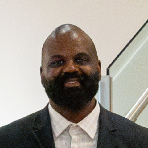 A headshot of a smiling Black man with a full beard, wearing a dark grey blazer and a white collared dress shirt against a minimalist white wall background.