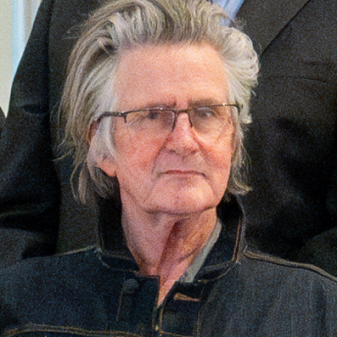 A close-up photograph shows an individual with messy, graying hair and glasses, wearing a dark denim jacket over a collared shirt.