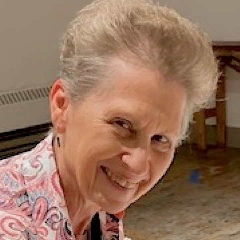 A smiling person with short gray hair tilts their head forward while wearing a pink and white patterned shirt.