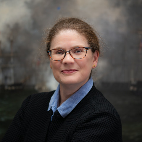 A headshot of a woman with brown hair tied back, wearing tortoiseshell glasses, a light blue collared shirt, and a black sweater.