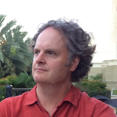 A man with wavy, dark hair and a receding hairline looks slightly to his left while wearing a red, collared button-down shirt outdoors.