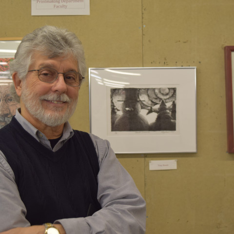 A smiling man with gray hair, a beard, and glasses stands with his arms crossed in front of a wall showcasing several framed art prints.