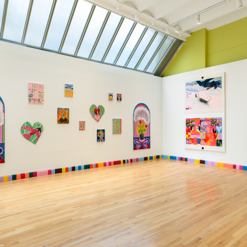 An art gallery filled with natural light from a large skylight showcases vibrant, colorful paintings displayed across stark white walls above a distinctly patterned, multi-colored baseboard and a light wood floor.