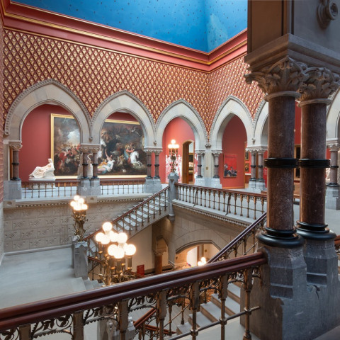 An ornate, Gothic Revival museum interior features patterned red walls, pointed stone arches, a bright blue ceiling, and a grand central staircase illuminated by spherical lamps.