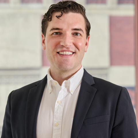 A smiling man with wavy dark hair wears a navy blue blazer over an unbuttoned white shirt, posing for a professional headshot in front of a window.