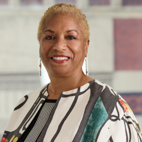 A smiling woman with short, blonde, curly hair wears long drop earrings and a white jacket adorned with a bold, colorful abstract pattern.