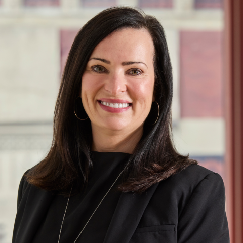 A professional portrait of a smiling woman with shoulder-length dark brown hair, wearing a black blazer, a black top, large hoop earrings, and a long silver pendant necklace against a softly blurred architectural background.