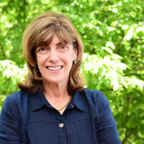 A woman with shoulder-length brown hair and bangs smiles directly at the camera while wearing a dark blue button-down shirt against a vibrant, blurry background of green leaves.