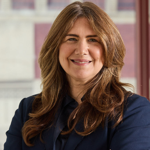 A professional portrait of a woman with long brown hair smiling with her arms crossed, wearing a dark blue suit blazer and a gold watch.
