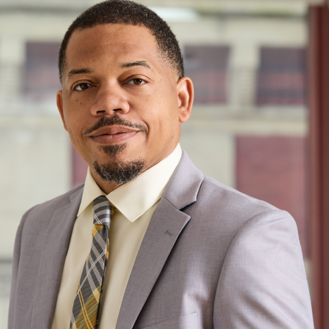 A headshot of a man with short dark hair, a mustache, and a goatee, wearing a light grey suit jacket, a cream-colored collared shirt, and a yellow, grey, and white plaid tie.